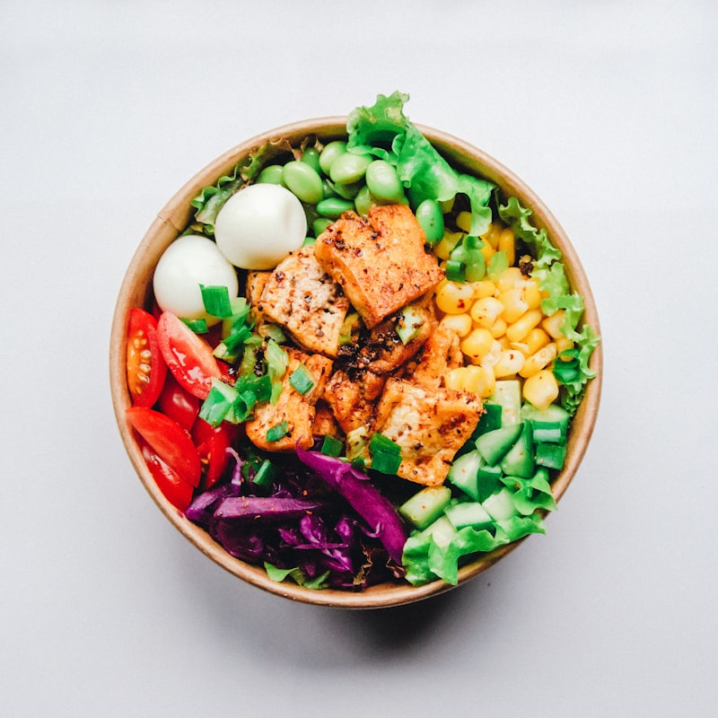 Wellness bowl with seared tofu, edamame, tomatoes, corn, and purple cabbage in a cardboard container